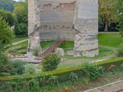 A tall, ancient cylindrical stone tower with brick accents and a modern metal staircase leading to its base, situated in a grassy park area next to railway tracks.