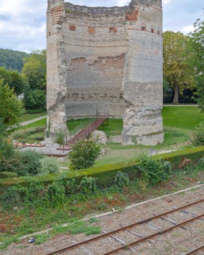 A tall, ancient cylindrical stone tower with brick accents and a modern metal staircase leading to its base, situated in a grassy park area next to railway tracks.