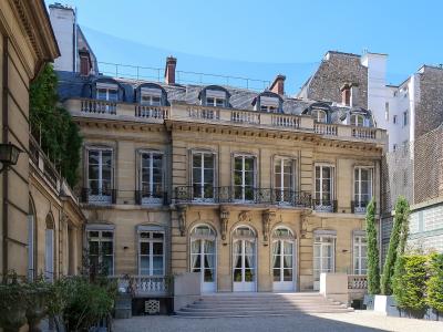 A beige stone private mansion with a gravel courtyard, arched ground-floor windows, a central balcony, and a mansard roof with dormer windows.