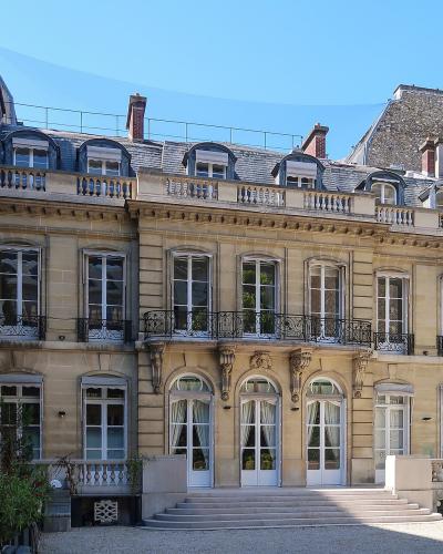 A beige stone private mansion with a gravel courtyard, arched ground-floor windows, a central balcony, and a mansard roof with dormer windows.