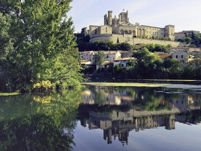 A large, fortified cathedral complex perched on a hill overlooking a river with lush green trees in the foreground and a clear reflection in the water.