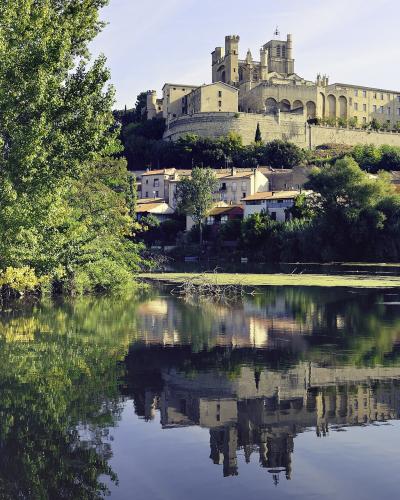 A large, fortified cathedral complex perched on a hill overlooking a river with lush green trees in the foreground and a clear reflection in the water.