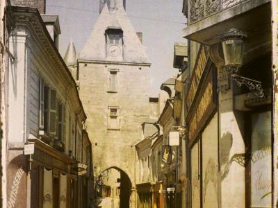 A vintage colorized photograph showing a narrow street leading toward a stone clock tower with an arched passage at its base, flanked by buildings with shop signs including "PATISSERIE" and "CONFISERIE," and a handwritten label at the bottom readi