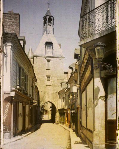 A vintage colorized photograph showing a narrow street leading toward a stone clock tower with an arched passage at its base, flanked by buildings with shop signs including "PATISSERIE" and "CONFISERIE," and a handwritten label at the bottom readi