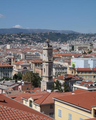 A high-angle view of the historic old town of Nice, featuring terracotta rooftops, a prominent clock tower (the Église Saint-Nicolas), and distant hills under a clear blue sky.