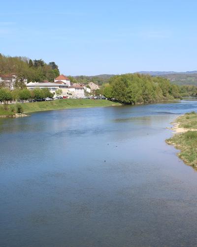 A wide river flowing through a green valley with a town on the left bank and hills in the distance under a blue sky.