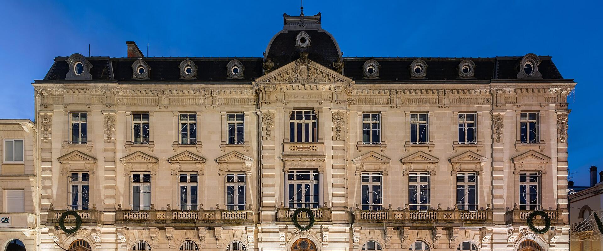 A grand, ornate neoclassical building at dusk, featuring a central pediment, dormer windows, balconies with wreaths, and the word “HÔTEL DE VILLE” above the main entrance; bicycles are parked in front on a paved plaza.