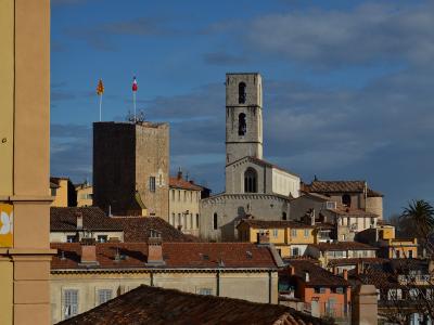 A view of the old town of Grasse, France, featuring the stone bell tower and facade of the Cathédrale Notre-Dame du Puy alongside the Tour du Palais (a fortified tower with French and Provençal flags) against a blue sky.