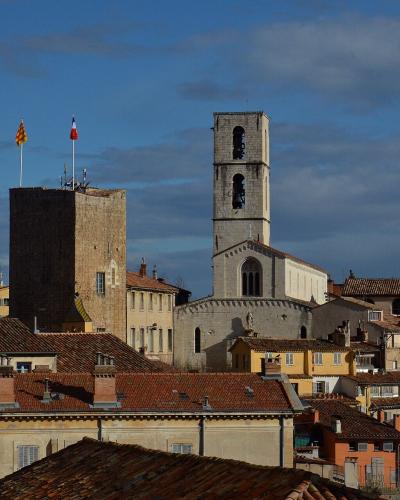 A view of the old town of Grasse, France, featuring the stone bell tower and facade of the Cathédrale Notre-Dame du Puy alongside the Tour du Palais (a fortified tower with French and Provençal flags) against a blue sky.