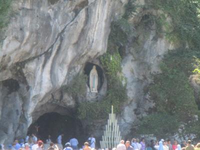 A limestone cliff face with a natural cave opening at the base and a statue of the Virgin Mary set into a niche above it, surrounded by greenery, with a crowd of visitors gathered behind a railing in the foreground.