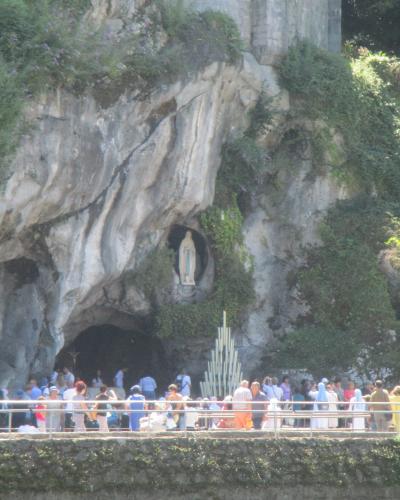 A limestone cliff face with a natural cave opening at the base and a statue of the Virgin Mary set into a niche above it, surrounded by greenery, with a crowd of visitors gathered behind a railing in the foreground.