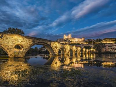 A long, illuminated medieval stone bridge with multiple arches spanning a river at twilight, with a large fortified cathedral and town buildings visible on the hill in the background.