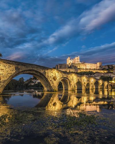 A long, illuminated medieval stone bridge with multiple arches spanning a river at twilight, with a large fortified cathedral and town buildings visible on the hill in the background.