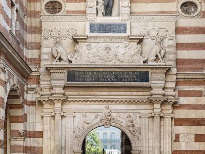 The image shows an ornate stone archway with a statue of a historical figure above it, set within a building featuring distinctive red and white striped brickwork, with Latin inscriptions on plaques.