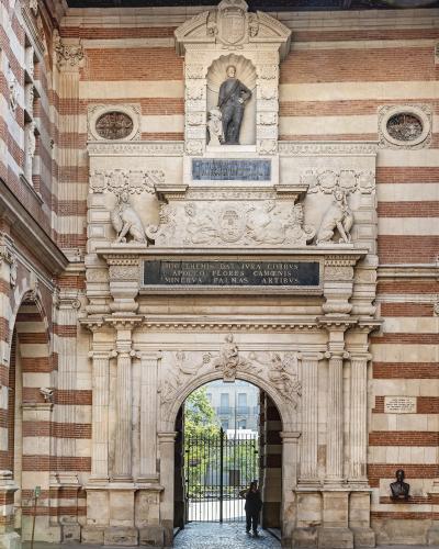 The image shows an ornate stone archway with a statue of a historical figure above it, set within a building featuring distinctive red and white striped brickwork, with Latin inscriptions on plaques.
