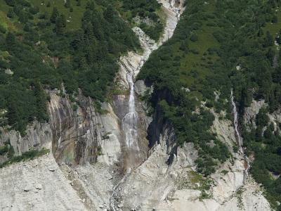 A high-angle view of a steep, rocky mountain valley with multiple waterfalls cascading down grey cliffs into a scree-covered basin below, surrounded by green alpine slopes and coniferous trees.