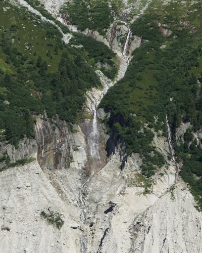 A high-angle view of a steep, rocky mountain valley with multiple waterfalls cascading down grey cliffs into a scree-covered basin below, surrounded by green alpine slopes and coniferous trees.