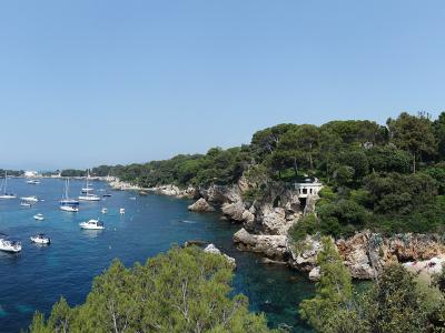A panoramic view of a sheltered bay with numerous sailboats anchored in clear blue water, bordered by a rocky coastline covered in dense green pine trees and a small white building perched on the cliffs.