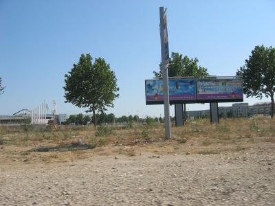 A vacant, gravelly lot with a large billboard advertising a future aquatic center ("Votre futur stade nautique") and a partially constructed or under-development sports complex visible in the background.