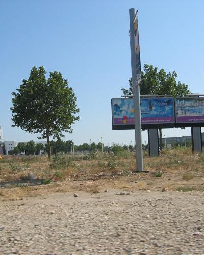 A vacant, gravelly lot with a large billboard advertising a future aquatic center ("Votre futur stade nautique") and a partially constructed or under-development sports complex visible in the background.