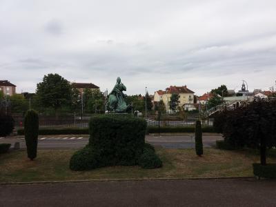A bronze statue of a seated female figure (likely representing the Republic) on a manicured hedge base, situated in a grassy median or park area next to a road with railway tracks and buildings in the background.