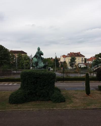 A bronze statue of a seated female figure (likely representing the Republic) on a manicured hedge base, situated in a grassy median or park area next to a road with railway tracks and buildings in the background.
