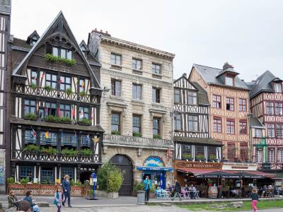 A row of historic half-timbered and stone buildings lining a paved public square, with outdoor café seating, pedestrians, and visible signage including “LES MARAICHERS” and “BAR LE CHATELET”.