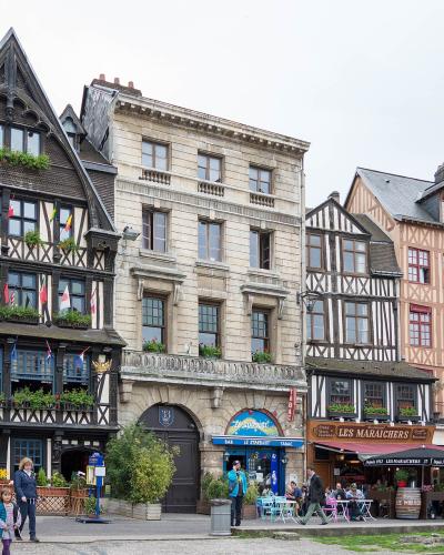 A row of historic half-timbered and stone buildings lining a paved public square, with outdoor café seating, pedestrians, and visible signage including “LES MARAICHERS” and “BAR LE CHATELET”.