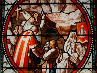 A stained-glass window depicting a religious scene with clergy, nuns, and a figure in a cave, featuring a red banner with "CREANTS LOURDES" text.