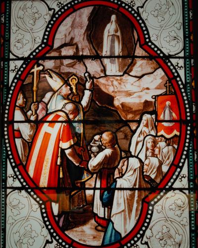 A stained-glass window depicting a religious scene with clergy, nuns, and a figure in a cave, featuring a red banner with "CREANTS LOURDES" text.