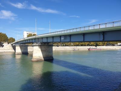 A modern steel girder bridge with stone piers spanning a wide river under a blue sky, with a barge visible on the water and trees lining the far bank; one pier bears an inscription reading "1931 - ANNI MCDXXXI".