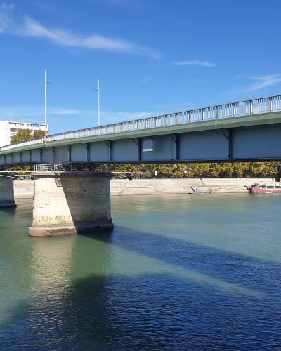 A modern steel girder bridge with stone piers spanning a wide river under a blue sky, with a barge visible on the water and trees lining the far bank; one pier bears an inscription reading "1931 - ANNI MCDXXXI".