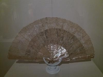 A close-up photograph of an antique lace fan displayed on a white pedestal inside a glass case, with the fan's sticks reflecting light.