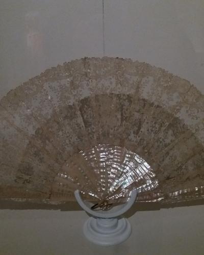 A close-up photograph of an antique lace fan displayed on a white pedestal inside a glass case, with the fan's sticks reflecting light.