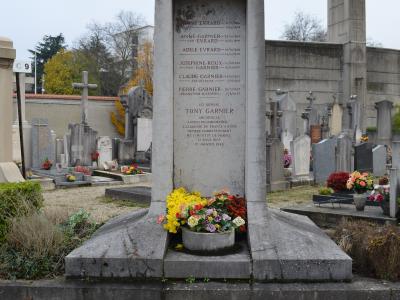 A stone tombstone in a cemetery, inscribed with "ICI REPOSE TONY GARNIER" and listing other family members, surrounded by flowers and other graves, with a modernist chapel tower visible in the background.
