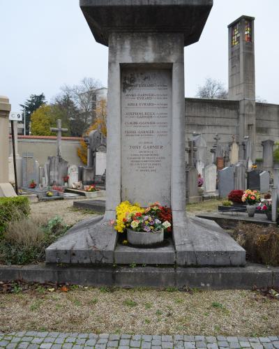 A stone tombstone in a cemetery, inscribed with "ICI REPOSE TONY GARNIER" and listing other family members, surrounded by flowers and other graves, with a modernist chapel tower visible in the background.