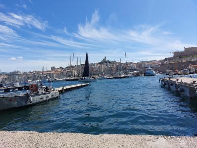 A sunny view of a busy harbor filled with sailboats and yachts, featuring a prominent stone fortification on the right and a hilltop basilica in the background.
