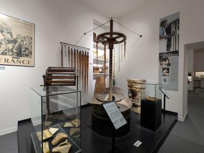 A museum gallery interior featuring ethnographic and archaeological artifacts, including a large wooden millstone mechanism, glass display cases with tools and objects, and wall panels with French text such as "FRANCE" and "La fabrique Macé".
