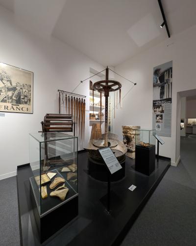A museum gallery interior featuring ethnographic and archaeological artifacts, including a large wooden millstone mechanism, glass display cases with tools and objects, and wall panels with French text such as "FRANCE" and "La fabrique Macé".