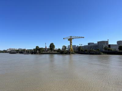 A wide river scene under a clear blue sky featuring a prominent yellow crane on a small island or peninsula, with modern buildings and other cranes visible in the background.