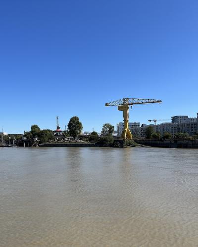 A wide river scene under a clear blue sky featuring a prominent yellow crane on a small island or peninsula, with modern buildings and other cranes visible in the background.