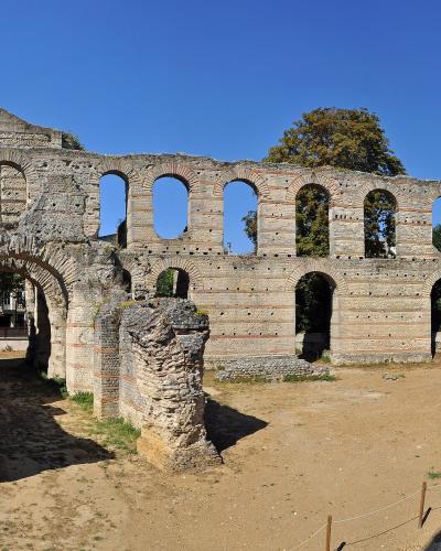 1. The image shows the ancient Roman ruins of the Amphitheatre of Bordeaux (Palais Gallien), featuring weathered stone walls with arched openings, set against a clear blue sky with modern buildings visible in the background.