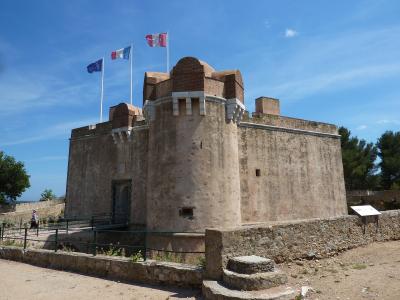 A stone fortress with a rounded tower, three flags (EU, French, and a red-and-white regional flag) flying above, under a blue sky, with a person walking near an entrance ramp.