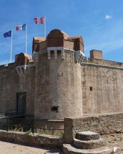 A stone fortress with a rounded tower, three flags (EU, French, and a red-and-white regional flag) flying above, under a blue sky, with a person walking near an entrance ramp.