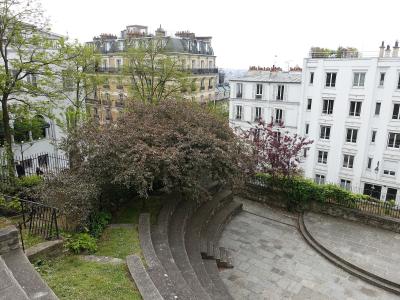 The image shows a small, semi-circular stone amphitheater or garden seating area surrounded by trees and typical Parisian apartment buildings with mansard roofs.