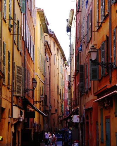 A narrow, sunlit cobblestone street lined with tall, ochre and terracotta-colored buildings featuring shuttered windows and vintage street lamps, with pedestrians walking in the distance.