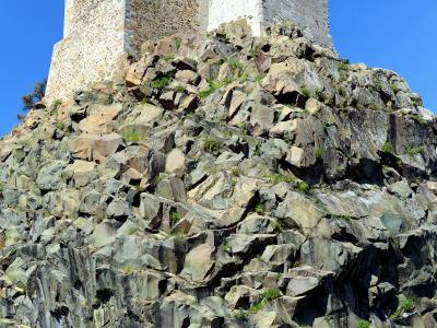 A low-angle photograph of a stone church with two distinct towers (one rougher stone, one smoother plaster) perched atop a steep, craggy rocky outcrop against a clear blue sky.
