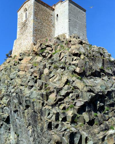 A low-angle photograph of a stone church with two distinct towers (one rougher stone, one smoother plaster) perched atop a steep, craggy rocky outcrop against a clear blue sky.
