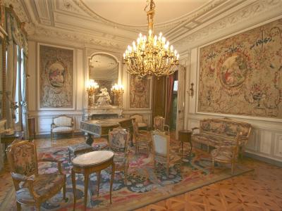An ornate 19th-century interior room featuring a large chandelier, gilded furniture, tapestries on the walls, a grand piano, and parquet flooring, identified as the music salon of the Musée Grobet-Labadié.