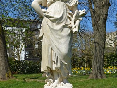 A white stone statue of a female figure in classical attire, standing on a rectangular pedestal in a grassy park with trees and yellow flowers in the background; no visible signage or text.